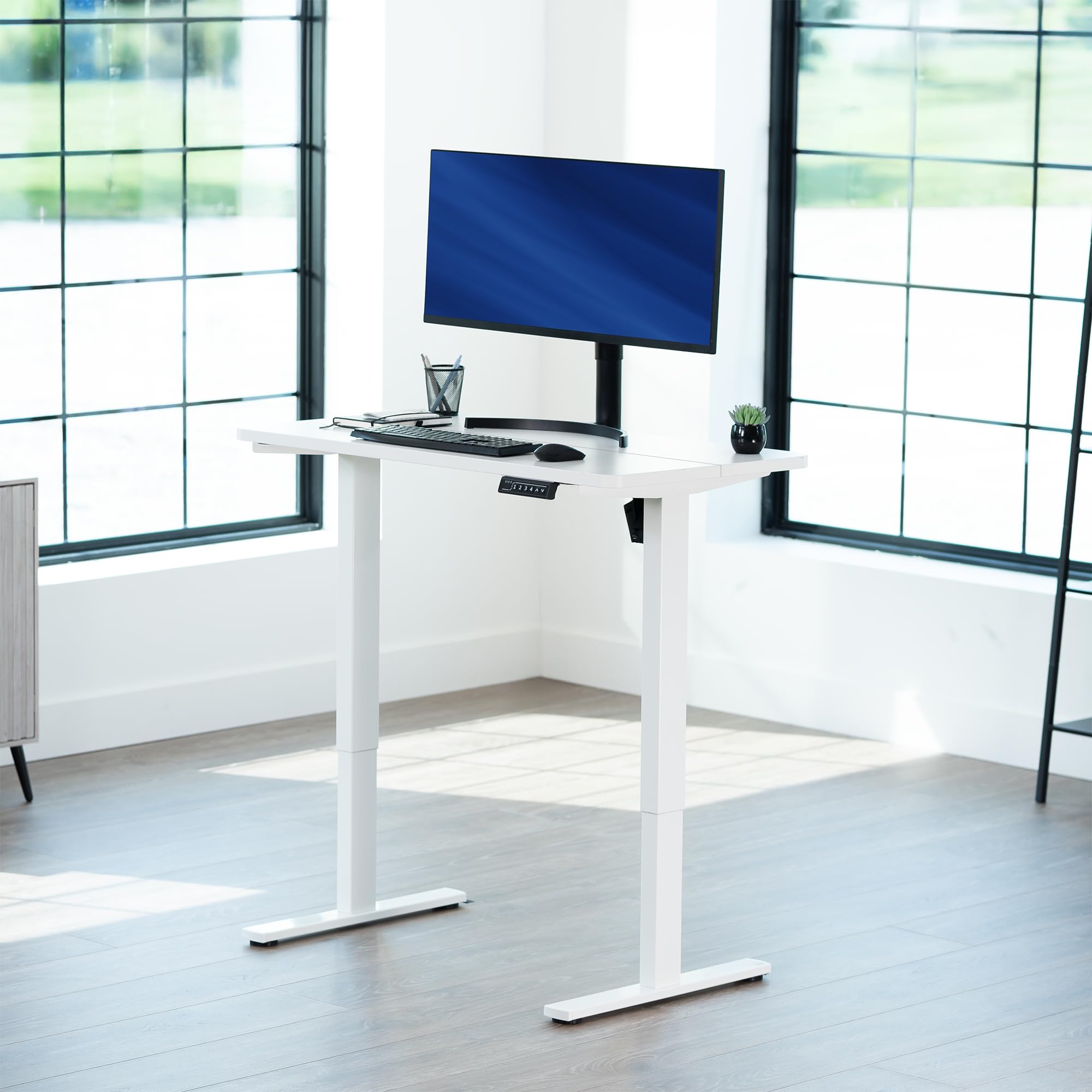 White standing desk with a computer setup in a bright room with large windows.
