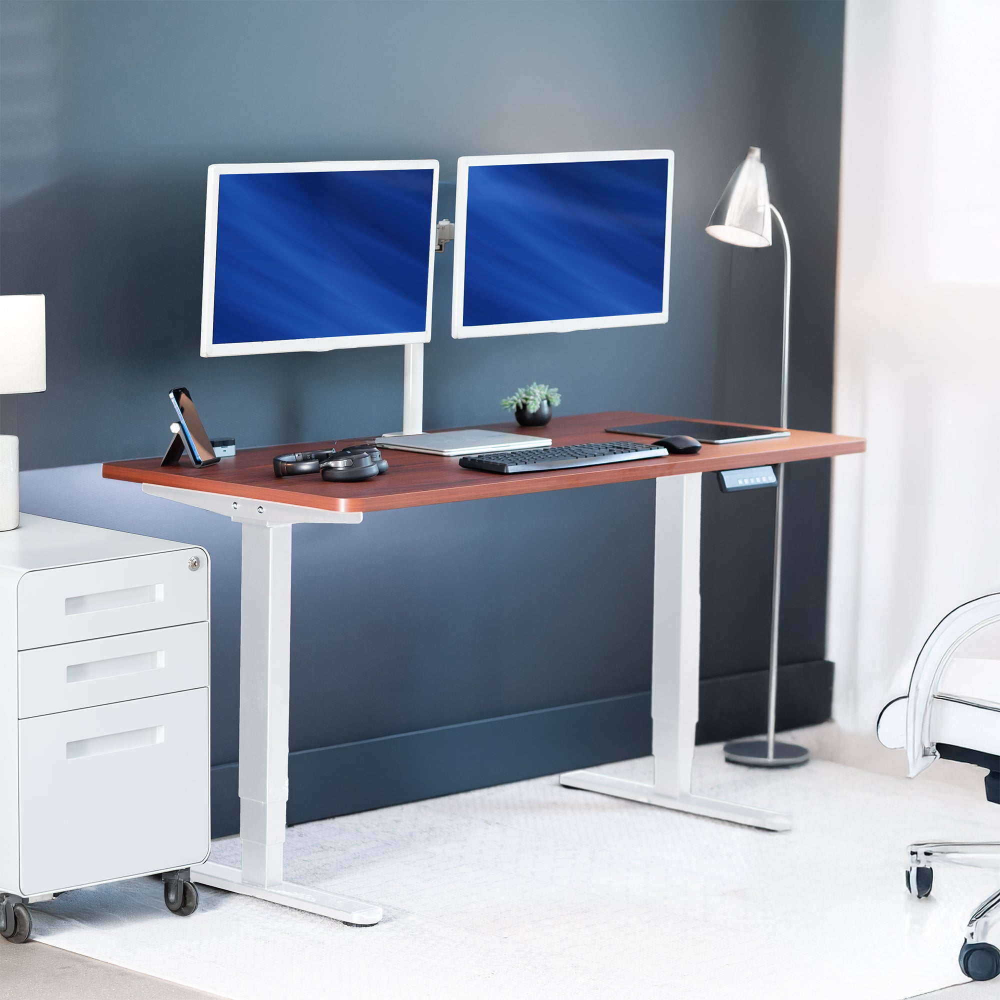 Modern office desk with two computer monitors, keyboard, and lamp against a gray wall.