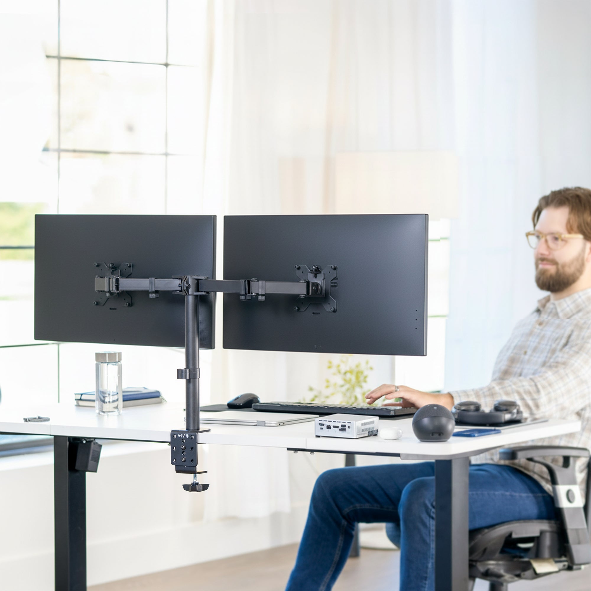 Person using a dual monitor stand at a desk in a bright room.