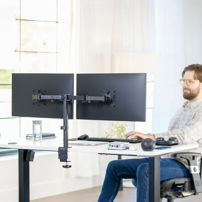 Person using a dual monitor stand at a desk in a bright room.
