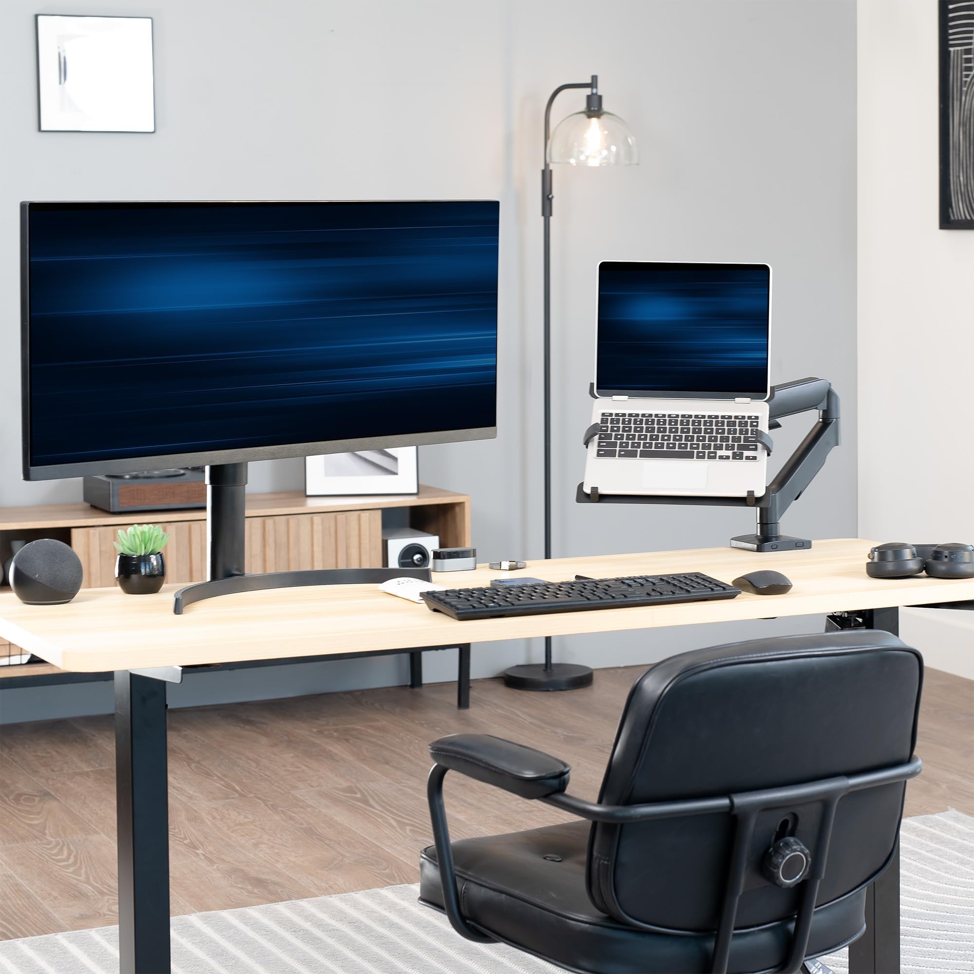 Modern office setup with a large freestanding monitor, laptop on an adjustable laptop mount, keyboard, and chair.