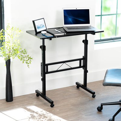Adjustable standing desk with laptop on a wooden floor near a window.