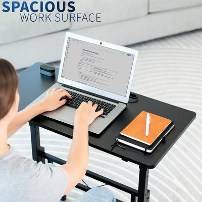 Person using a laptop on a black adjustable desk with a notebook and pen, against a light background.