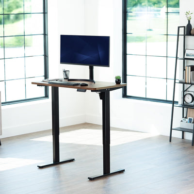 Rustic, adjustable height desk with a computer setup in a bright room with large windows.
