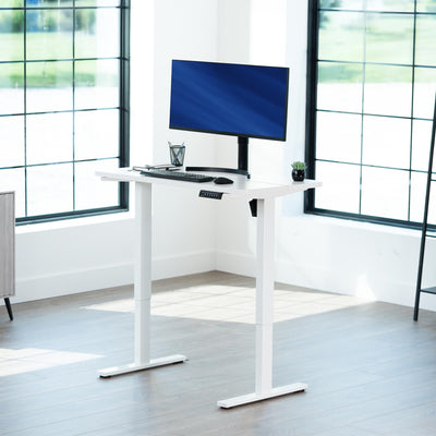 White standing desk with a computer setup in a bright room with large windows.