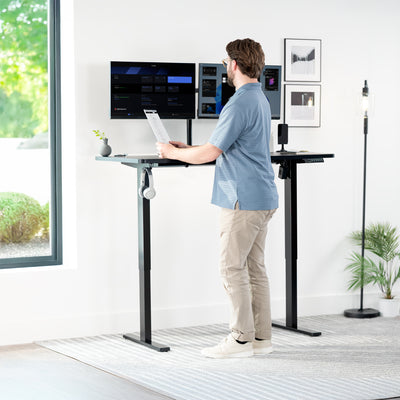 Person using a standing desk with multiple computer monitors in a bright room.
