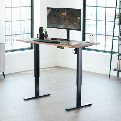 Adjustable height desk with computer setup in a bright room with large windows.