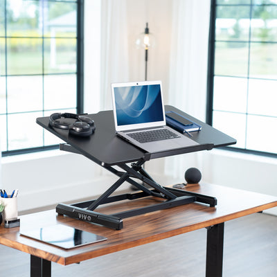 Black adjustable, tilting desk converter with a laptop on a wooden desk in a bright room.