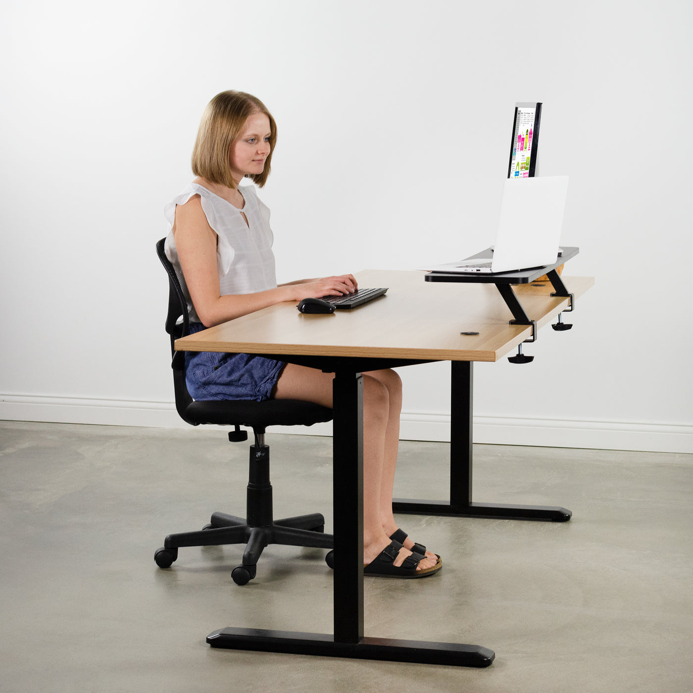 A woman working from an ergonomic office desk with a back of desk clamp on riser.